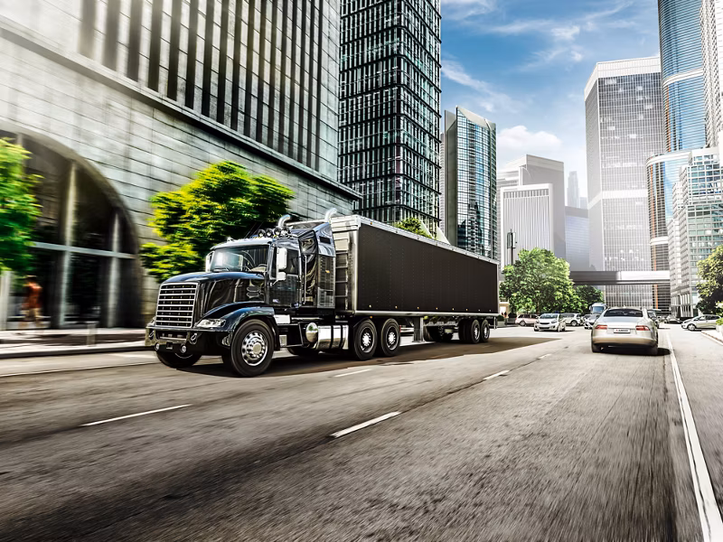 Large black heavy-duty truck driving on a multi-lane urban street surrounded by modern skyscrapers and glass buildings. The truck is hauling a long trailer, representing telematics and fleet management solutions for commercial vehicles. Bright daylight and city traffic create a professional logistics environment.
